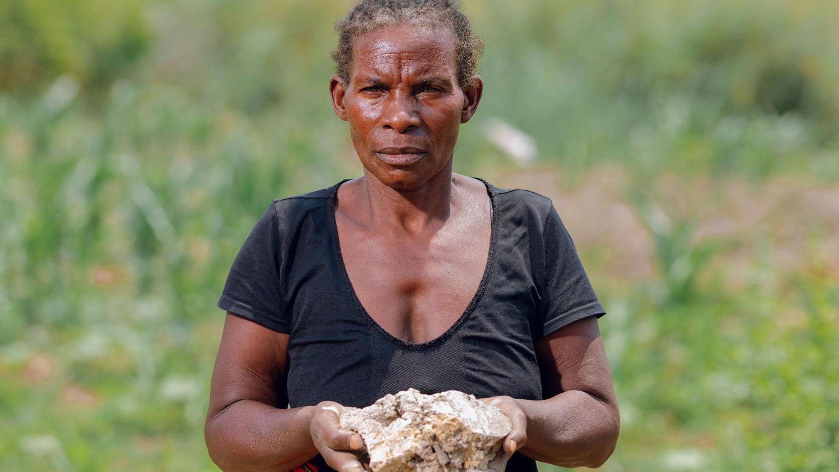 贊比亞農民瑪麗的照片肖像。她站在畫面中央，神情沉思，雙手舉著一大塊被污染的白色農作物土壤。她身後有綠意，她身穿黑色上衣和有圖案的裙子。