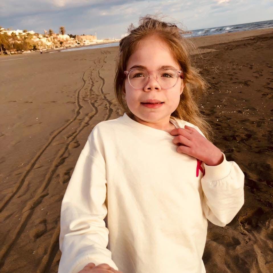 Sophia Scott está de pie en una playa. Es una niña pequeña de cabello largo y rubio, y lleva unas gafas grandes con montura rosa. Viste una sudadera de color crema y sonríe levemente a la cámara.