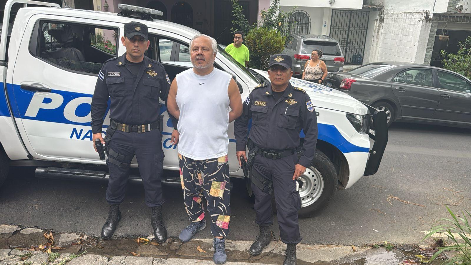 El abogado constitucionalista Enrique Anaya con los brazos hacia atrás y esposado, escoltado por dos policías delante de una camioneta policial.