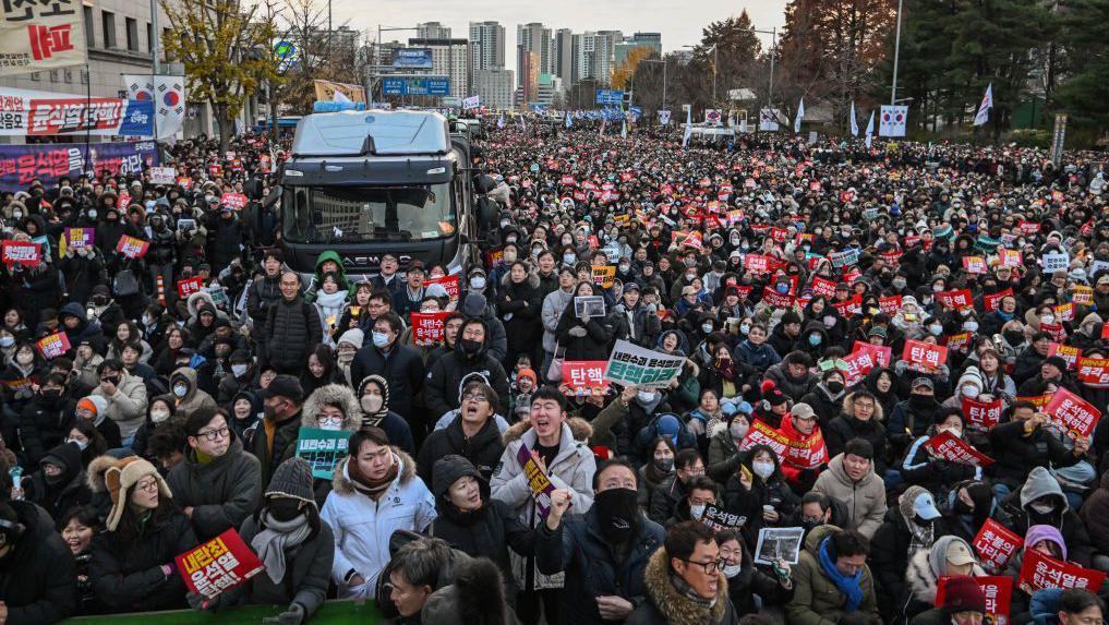 2024年12月7日，人們在首爾國會外參加示威活動，要求罷免韓國總統尹錫悅。大量身著冬裝的人群手持海報，揮舞著韓國國旗。中間是一輛黑色貨車，可能是警車。