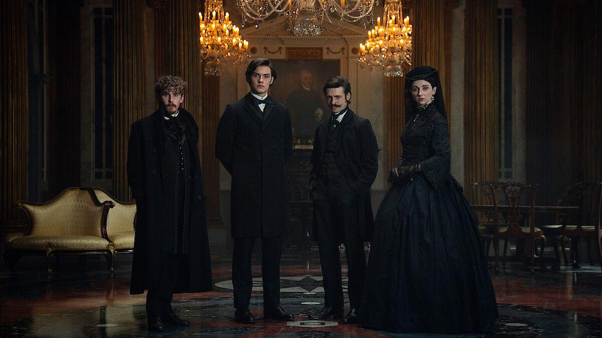 En un salón de estilo gótico o victoriano iluminado por grandes candelabros, cuatro personas posan de pie luciendo elegantes trajes de época negros.