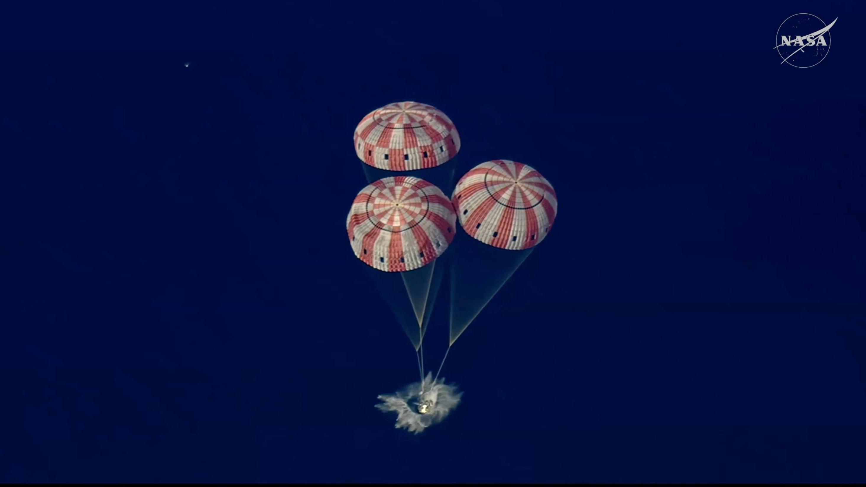 Imagen aérea capta tres paracaídas de rayas blancas y rojas infladas amortiguan el amerizaje de la cápsula que salpica al tocar el agua azul oscura del mar Pacífico