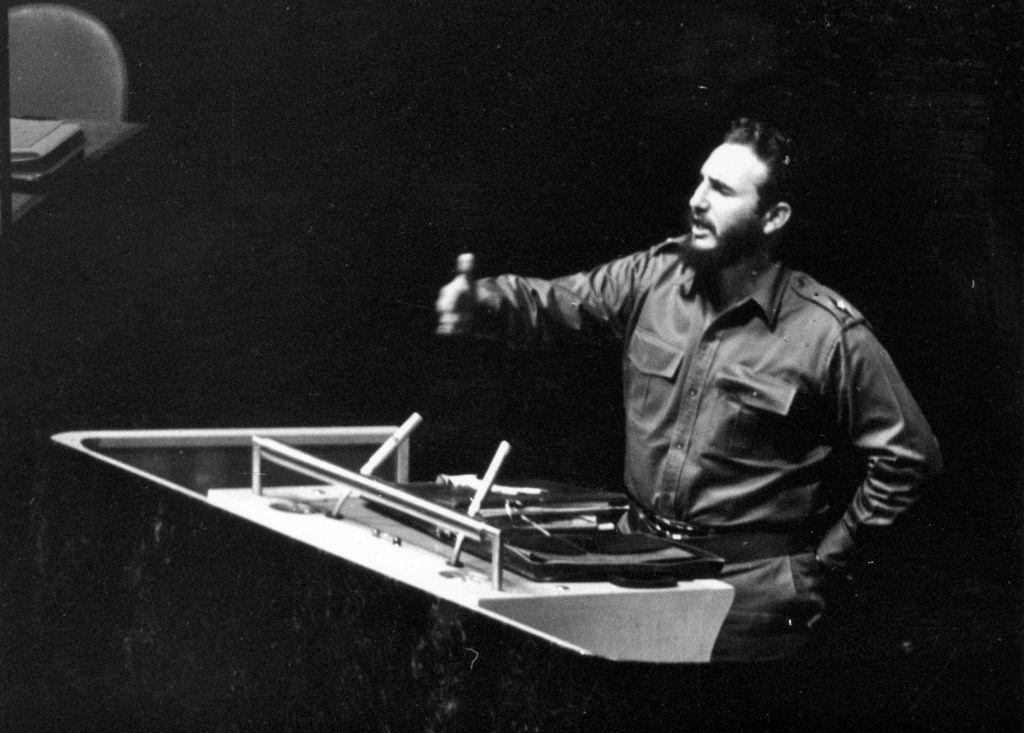 Imagen en blanco y negro de Fidel Castro hablando de pie desde el podio de oradores de la Asamblea General. Vestido con lo que parece un uniforme militar, Castro tiene su mano izquierda apoyada sobre la cintura y la derecha levantada en el aire mientras habla.