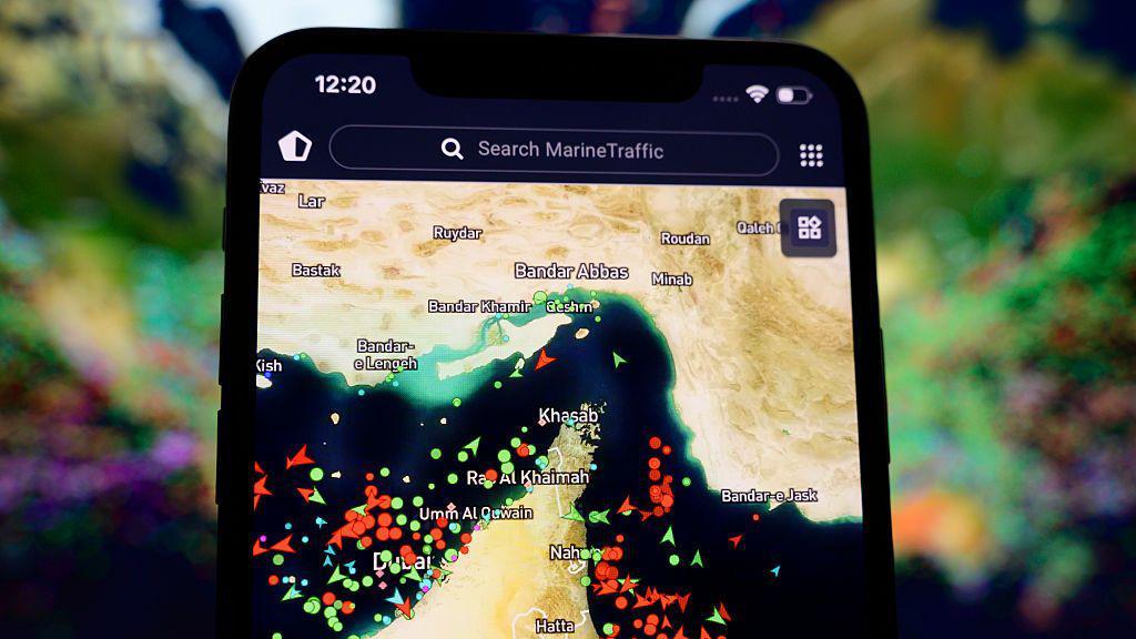 A tela de um smartphone exibe um mapa de rastreamento mar&iacute;timo. O mapa destaca uma regi&atilde;o do Golfo P&eacute;rsico e do Estreito de Ormuz, com diversas embarca&ccedil;&otilde;es representadas por pontos e &iacute;cones coloridos espalhados pelas rotas mar&iacute;timas. Nomes de cidades costeiras e &aacute;reas pr&oacute;ximas aparecem identificados no mapa, enquanto a barra de busca do aplicativo est&aacute; vis&iacute;vel na parte superior da tela. O fundo desfocado atr&aacute;s do celular sugere um ambiente externo com vegeta&ccedil;&atilde;o, mantendo o foco principal na visualiza&ccedil;&atilde;o do tr&aacute;fego naval.