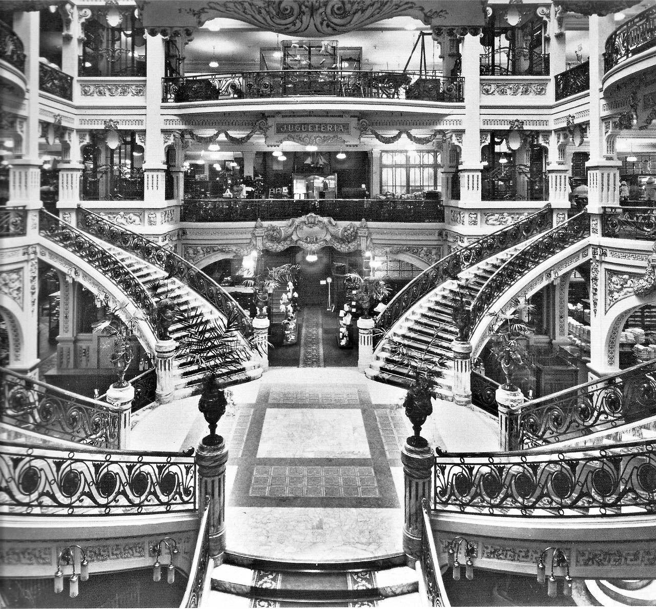 A imagem em preto e branco mostra o interior de uma grande loja de departamentos do in&iacute;cio do s&eacute;culo 20. O espa&ccedil;o &eacute; marcado por escadarias sim&eacute;tricas, v&aacute;rios andares com varandas ornamentadas, colunas decorativas e ilumina&ccedil;&atilde;o suspensa. Ao centro, h&aacute; um amplo v&atilde;o de circula&ccedil;&atilde;o, enquanto diferentes setores da loja podem ser vistos ao fundo, evidenciando a arquitetura sofisticada e o car&aacute;ter luxuoso do estabelecimento.