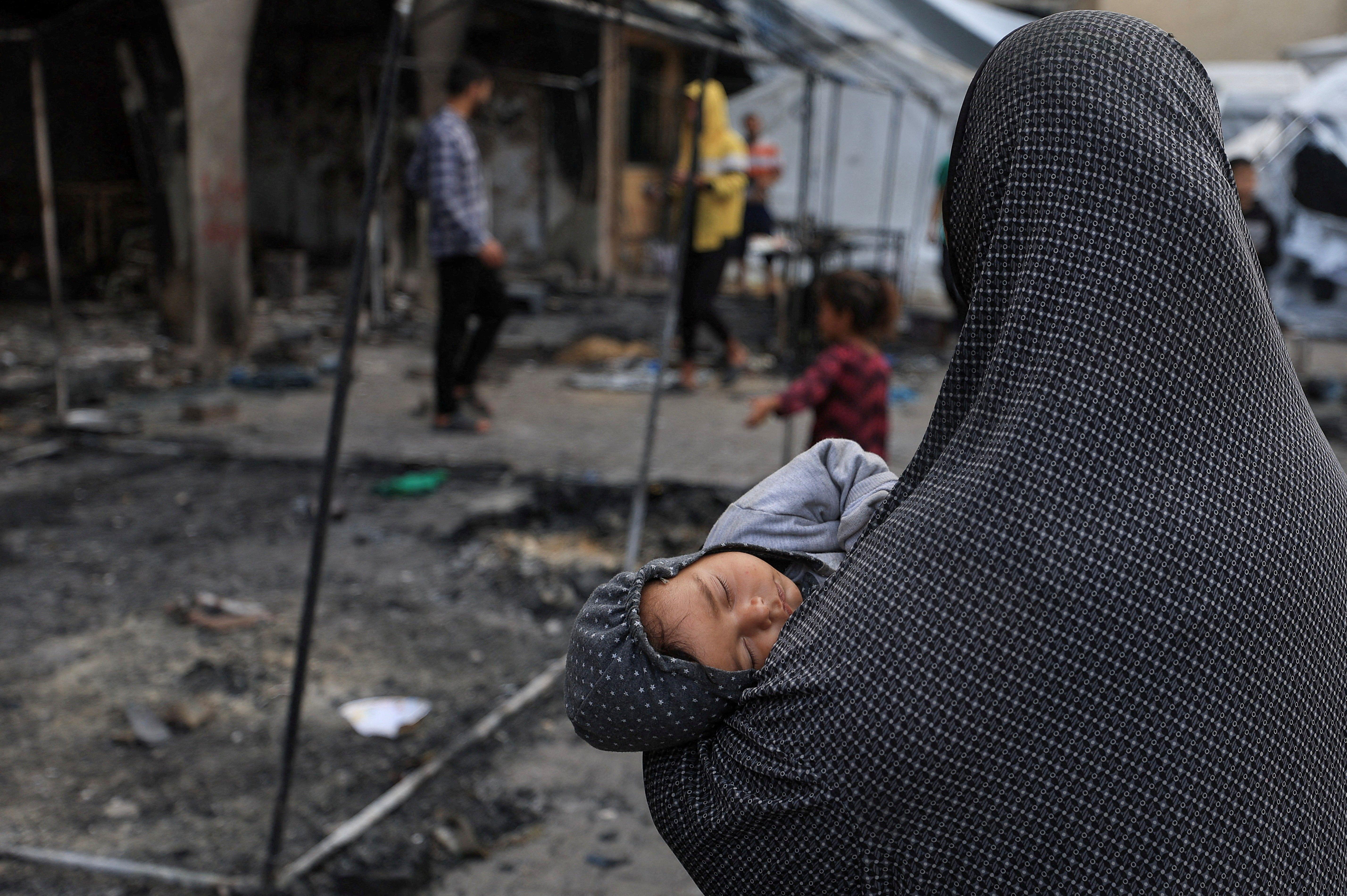 امرأة فلسطينية تحمل طفلاً، بينما يتفقد فلسطينيون الأضرار التي لحقت بمدرسة تؤوي النازحين، بعد غارة إسرائيلية، في مدينة غزة، 23 أبريل/نيسان 2025.