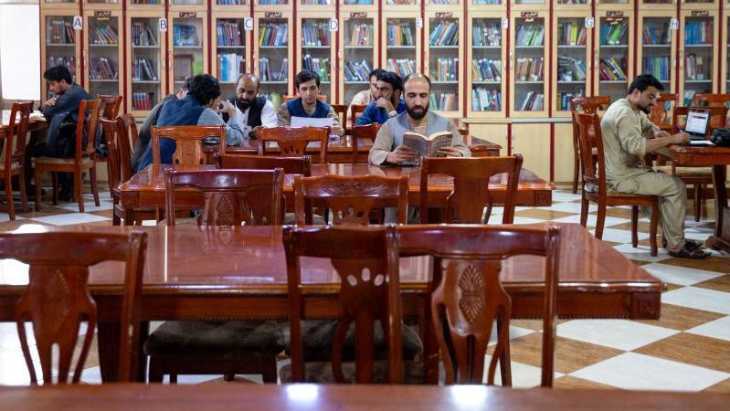 Los hombres se sientan en mesas en una habitación con estanterías al fondo.