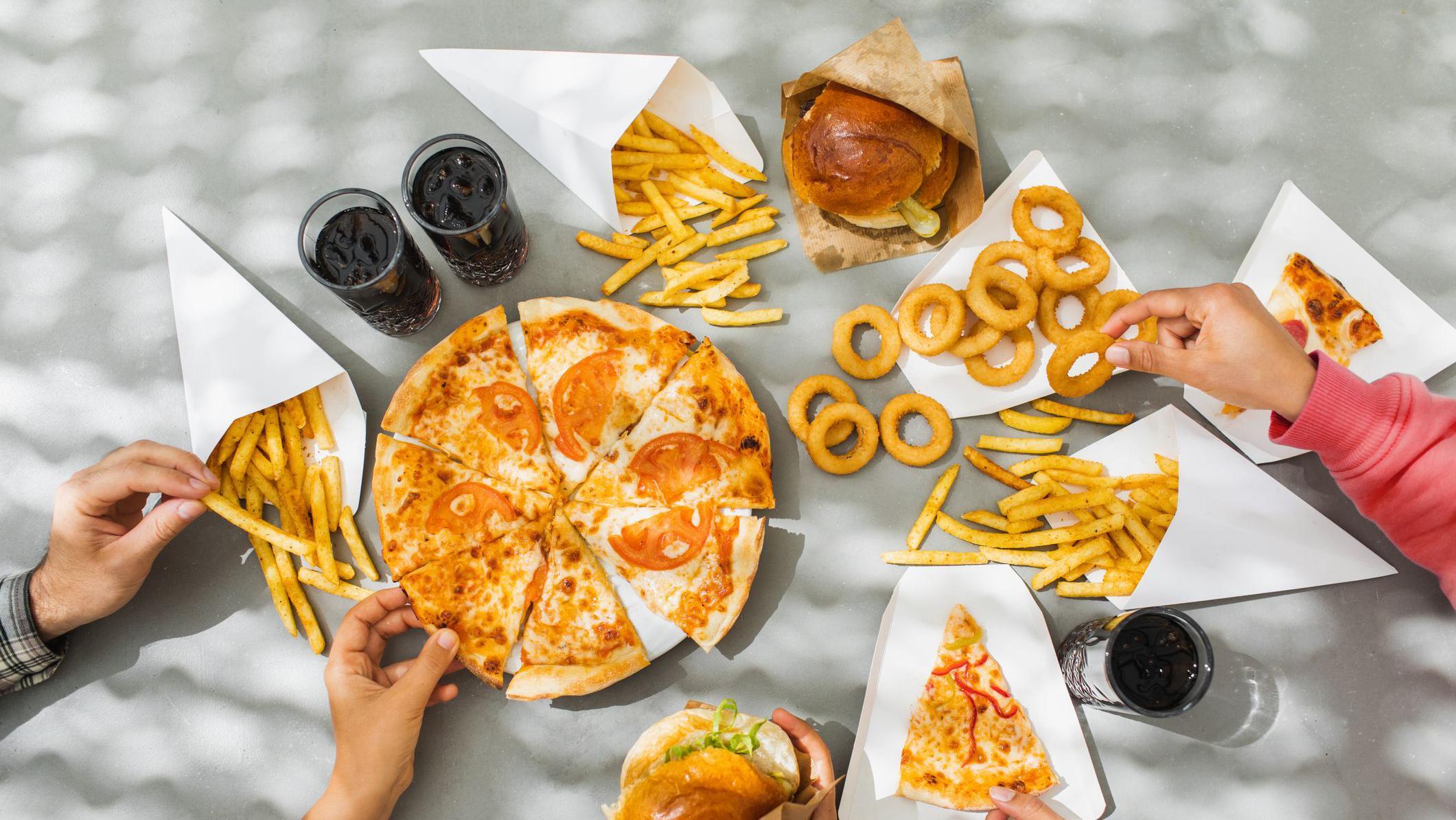 Una mesa llena de platos de pizza, hamburguesas en panecillos, aros de cebolla, papas fritas y refrescos de cola, todos ellos ejemplos de alimentos ultraprocesados. Varias manos se extienden y toman los diferentes alimentos.