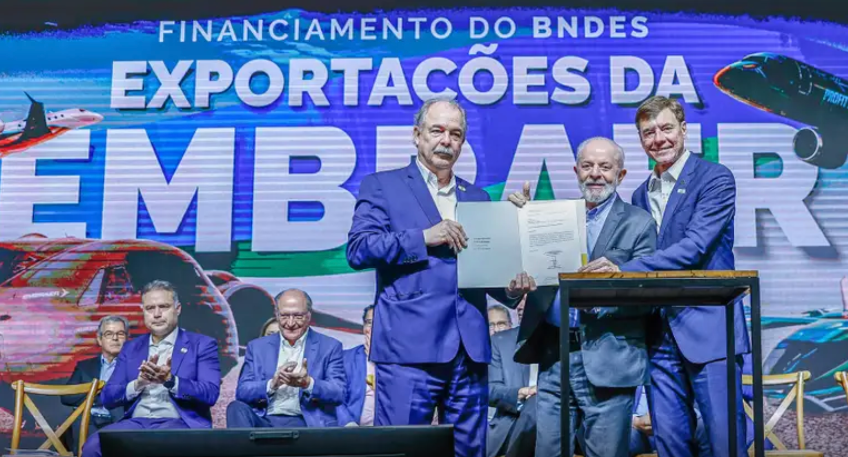 Políticos brasileiros, dentre eles o presidente Lula e o presidente do BNDES Aloizio Mercadante, na frente de um anúncio com o texto 