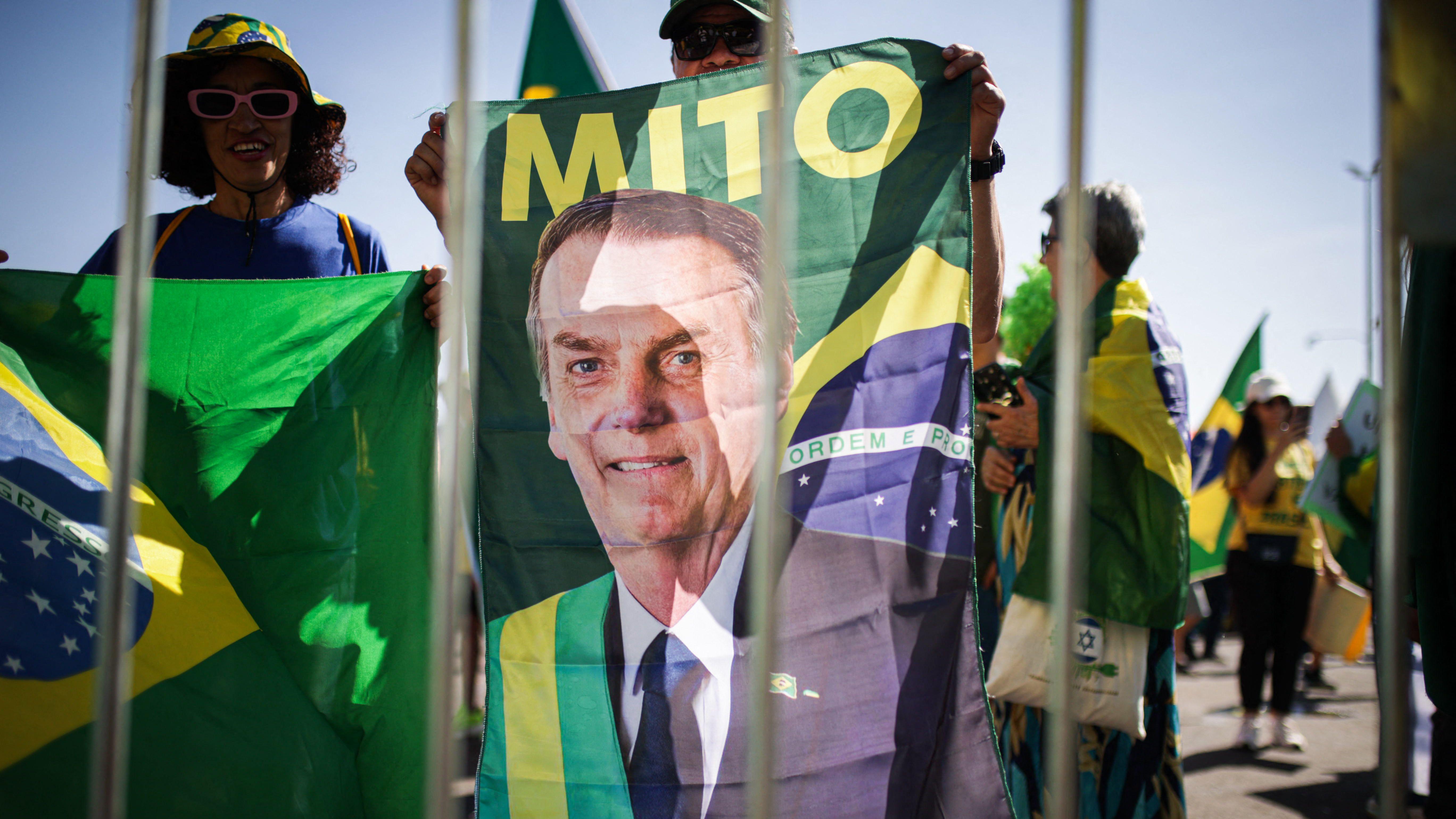 Manifestantes seguram bandeiras do Brasil e um grande estandarte com a imagem do ex-presidente Jair Bolsonaro durante manifesta&ccedil;&atilde;o. A cena &eacute; vista atrav&eacute;s de grades met&aacute;licas em primeiro plano, enquanto participantes vestindo roupas nas cores verde e amarelo se concentram em uma &aacute;rea aberta sob a luz do dia.