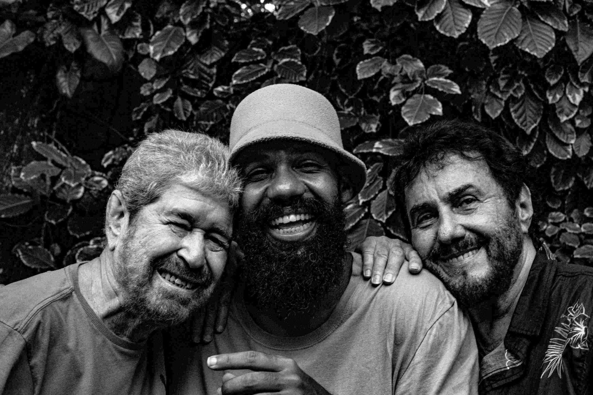 Dupla sorrindo para a foto com Russo Passapusso; eles estão próximos, abraçados, em frente a muro com plantas