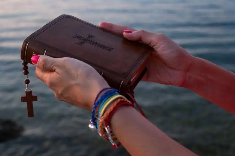 Las manos de una mujer con manillas de los colores de la bandera gay cargan una biblia, de la cual cuelga un rosario. 