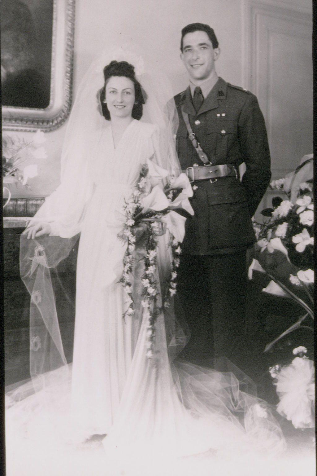 Elizabeth con vestido de novia, largo velo y ramo de flores grande; Robert con uniforme militar