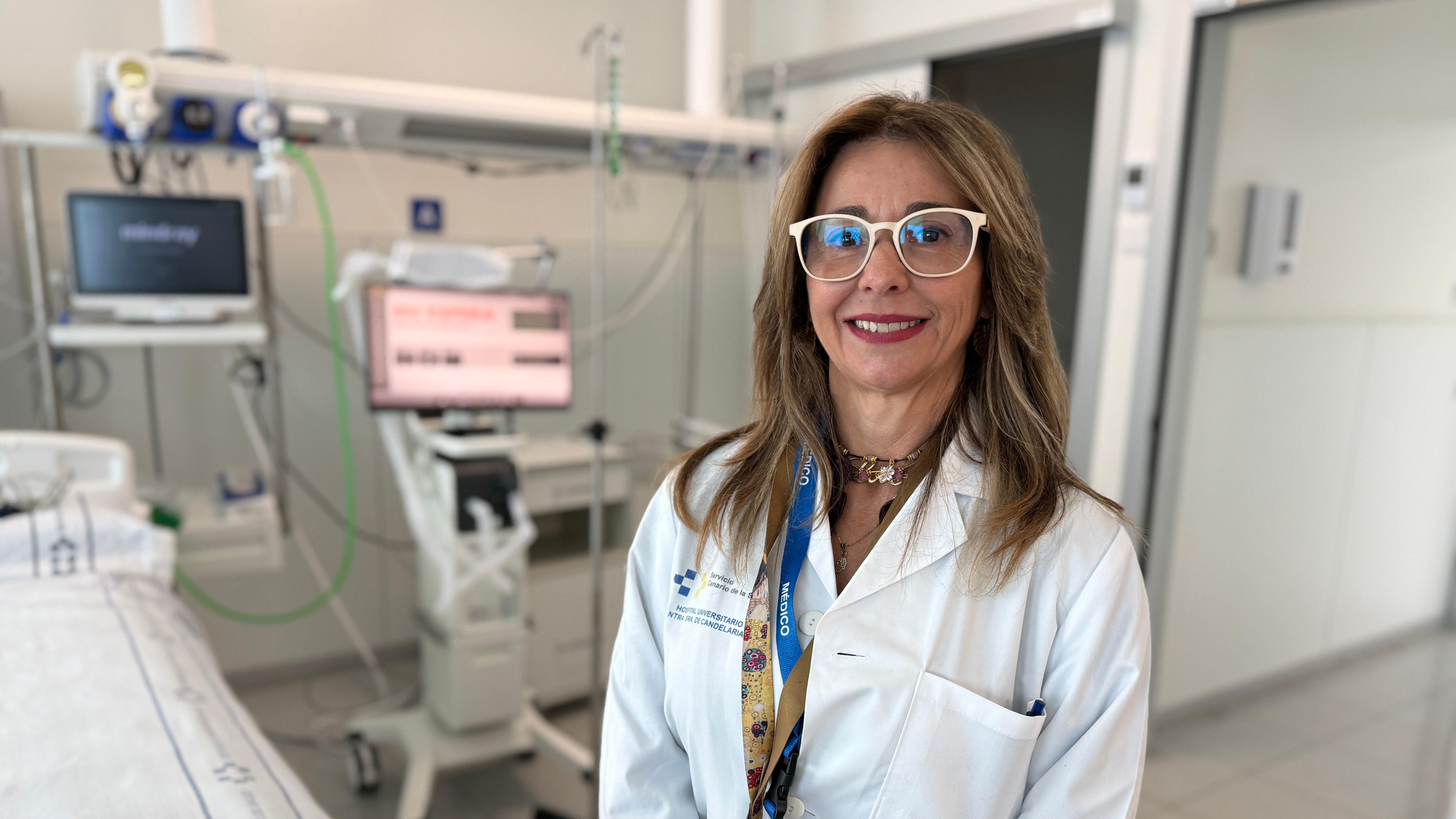 Una doctora con gafas blancas y el cabello largo y castaño sonríe. Se encuentra en una instalación médica con una cama hospitalaria a su lado y dos pantallas con varios cables alrededor detrás de ella.