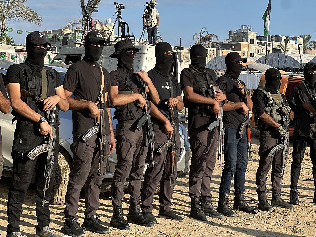 Una fila de 8 hombres, uno al lado de otro, todos con botas militares, vestidos con ropa oscura, pasamontañas que impide que se les vea la cara y gorra. Todos llevan en sus manos un arma larga tipo rifle. Están al aire libre. Al fondo se ven unas furgonetas y, sobre ellas, tres cámaras de video. Junto a una de ellas, un hombre.