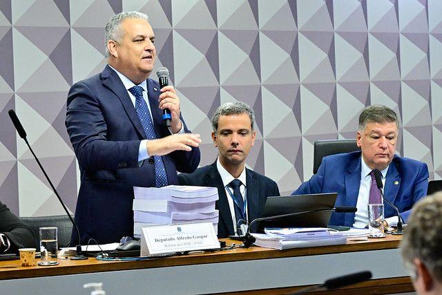 Gaspar fala ao microfone, de p&eacute;, com uma grande pilha de pap&eacute;is a sua frente, seu relat&oacute;rio para a CPMI 