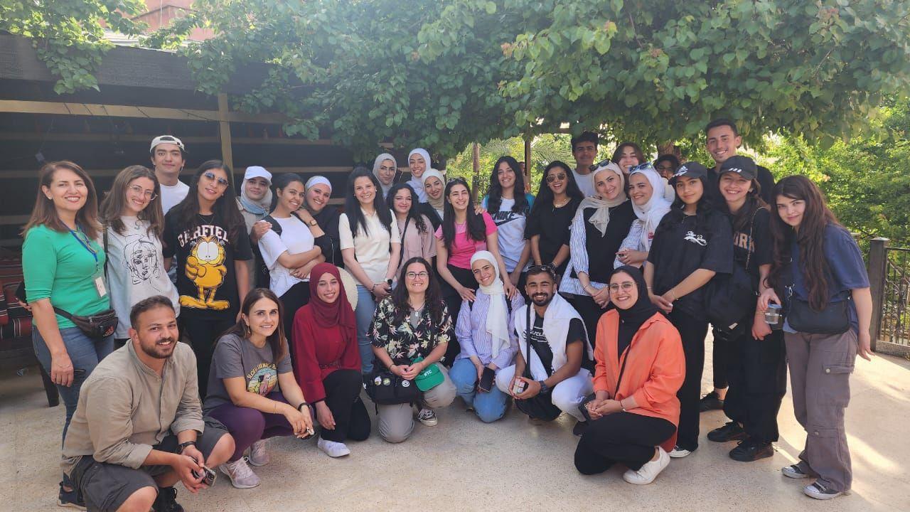 تُظهر الصورة مجموعة كبيرة من الشباب والشابات في صورة جماعية مبهجة في الهواء الطلق. يقف الأفراد في صفين، بعضهم يقف والآخر يجلس على الأرض، وكلهم يبتسمون للكاميرا. تتوسطهم الدكتورة دانة قاقيش.