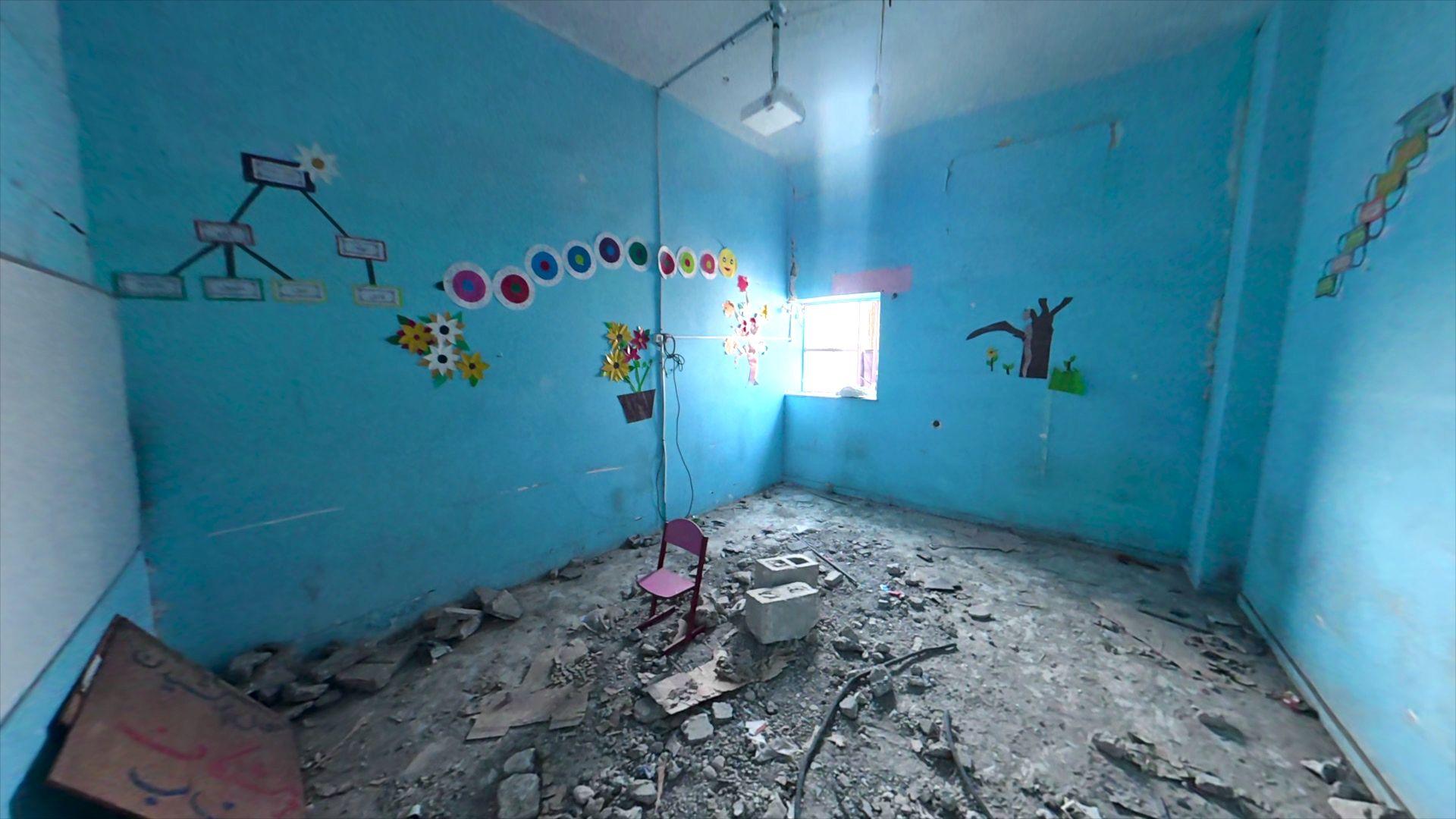 Interior de uma sala de aula com as paredes pintadas de azul-claro. Imagens de uma &aacute;rvore, flores e c&iacute;rculos coloridos decoram as paredes. Escombros cobrem o ch&atilde;o, com uma pequena cadeira rosa e dois blocos de concreto no meio da sala