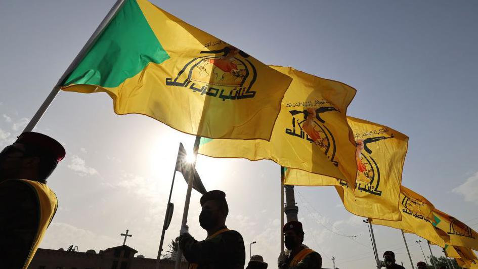 أشخاص ملثمون يحملون رايات صفراء وخضراء مكتوب عليها "كتائب حزب الله"، يسيرون في شوارع بغداد خلال جنازة أحد قادة الفصيل 