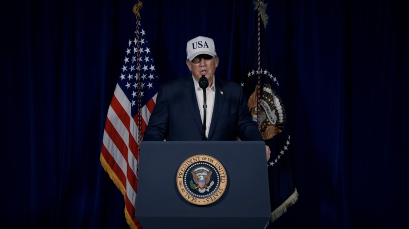 Donald Trump, vestido con una camisa blanca y un traje oscuro, tiene puesta una gorra que dice USA, habla desde el podio del presidente de Estados Unidos y atrás tiene la bandera de EE.UU.