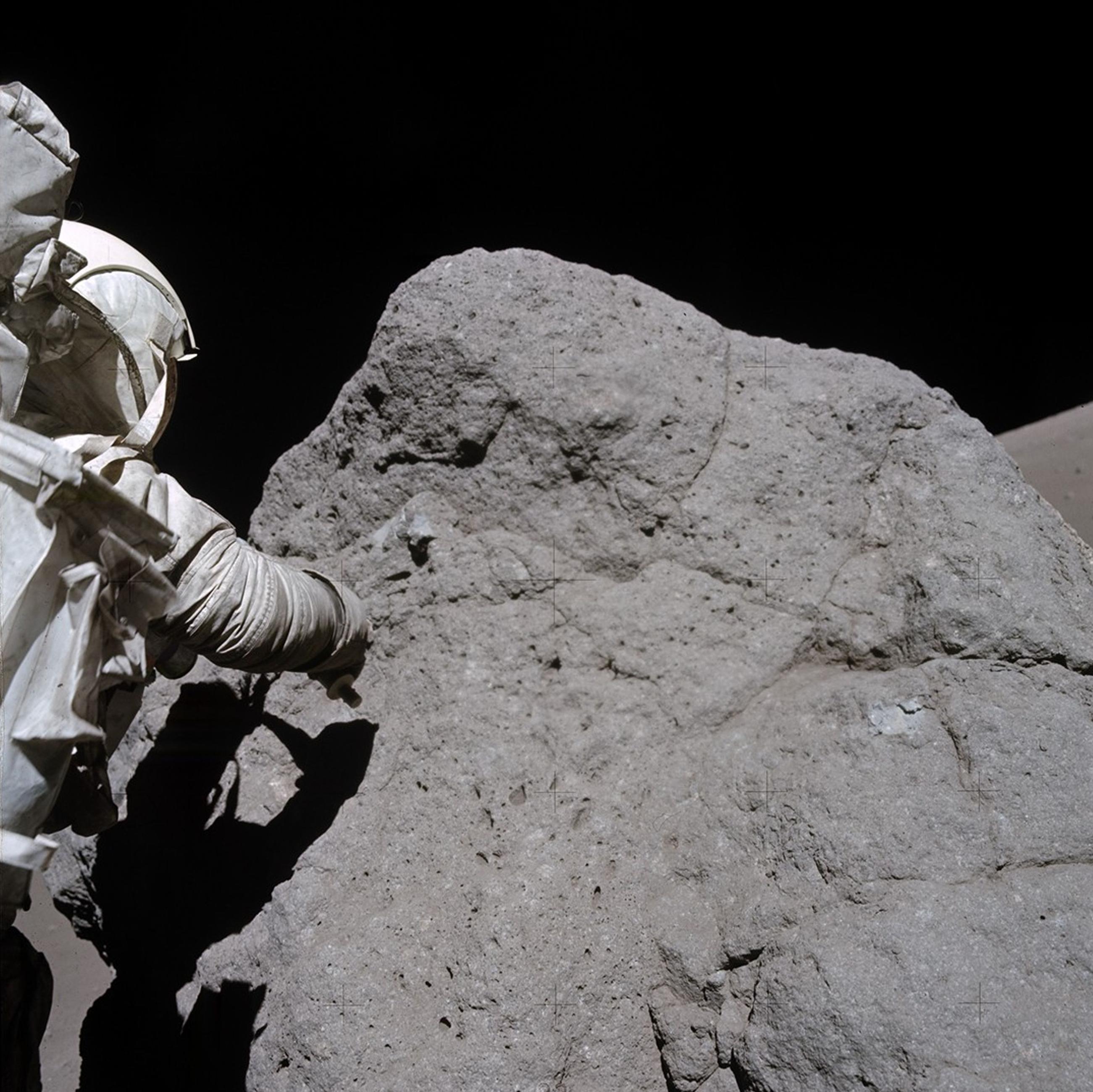O astronauta Harrison Schmitt, da Apollo 17, coleta amostras de rochas da Lua. A rocha &eacute; um pouco mais alta que ele e tem cerca de tr&ecirc;s vezes sua largura. Ele est&aacute; &agrave; esquerda da foto, de costas para a c&acirc;mera, vestindo um traje espacial branco da Nasa, e tenta coletar a amostra usando seu bra&ccedil;o direito. Seu corpo cria uma sombra sobre a grande rocha cinza escura.  