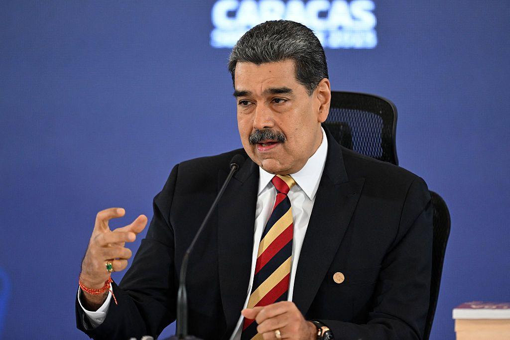 Nicolás Maduro, con traje oscuro y corbata de rayas, comparece ante un micr´fono con la mano derecha en alto.