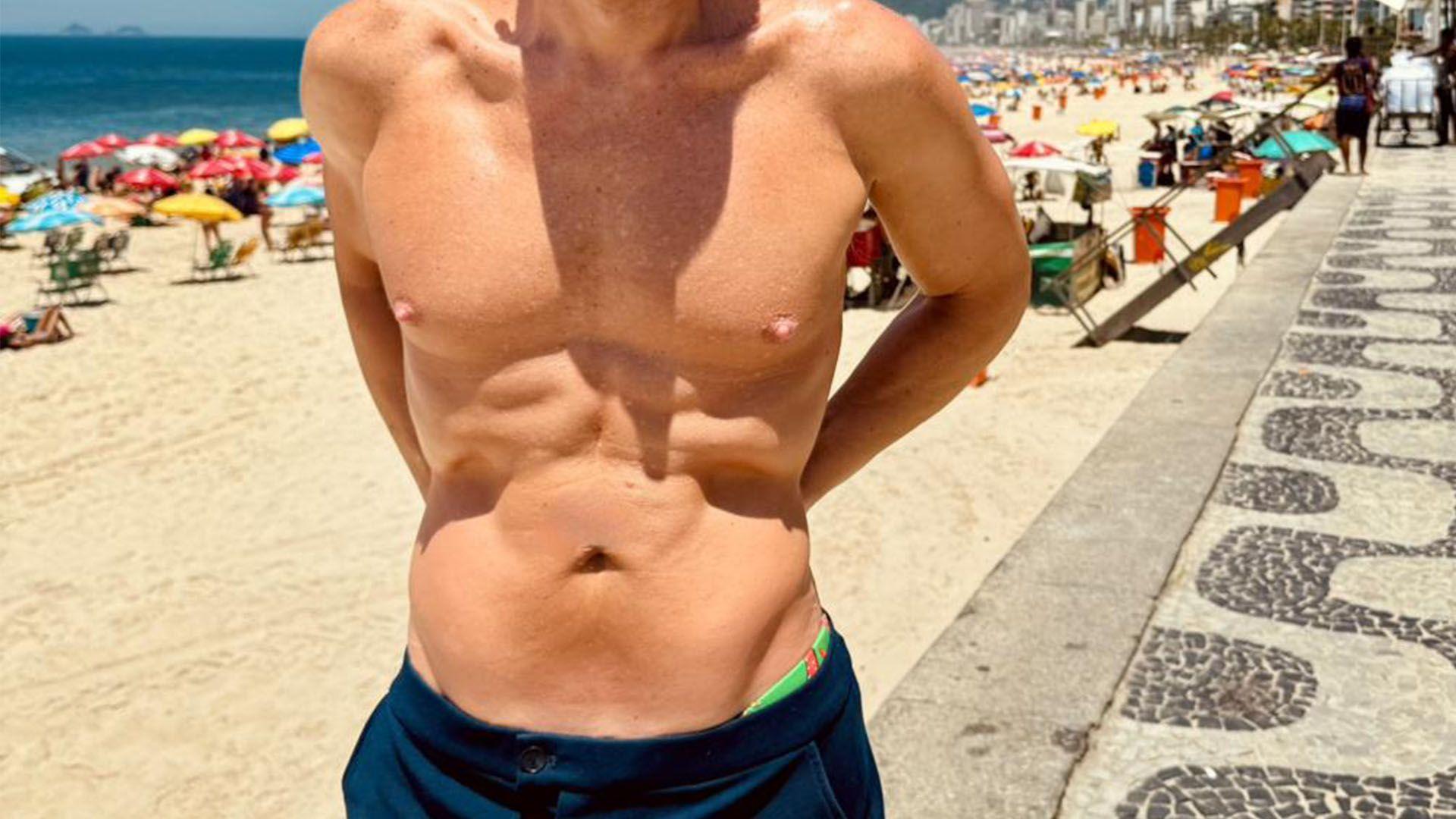 Foto de um homem sem rosto, com o torso musculoso à mostra na praia, usando shorts azuis. 