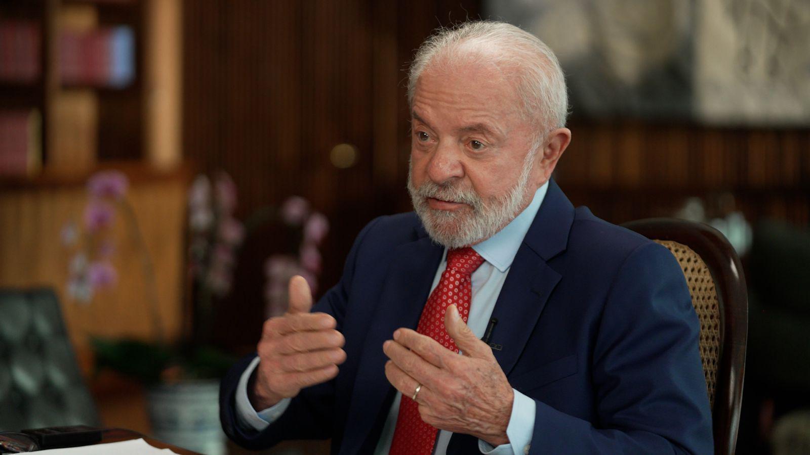 Lula viste una chaqueta de traje azul oscura, una camisa clara y una corbata roja. Está sentado en una silla de madera.
