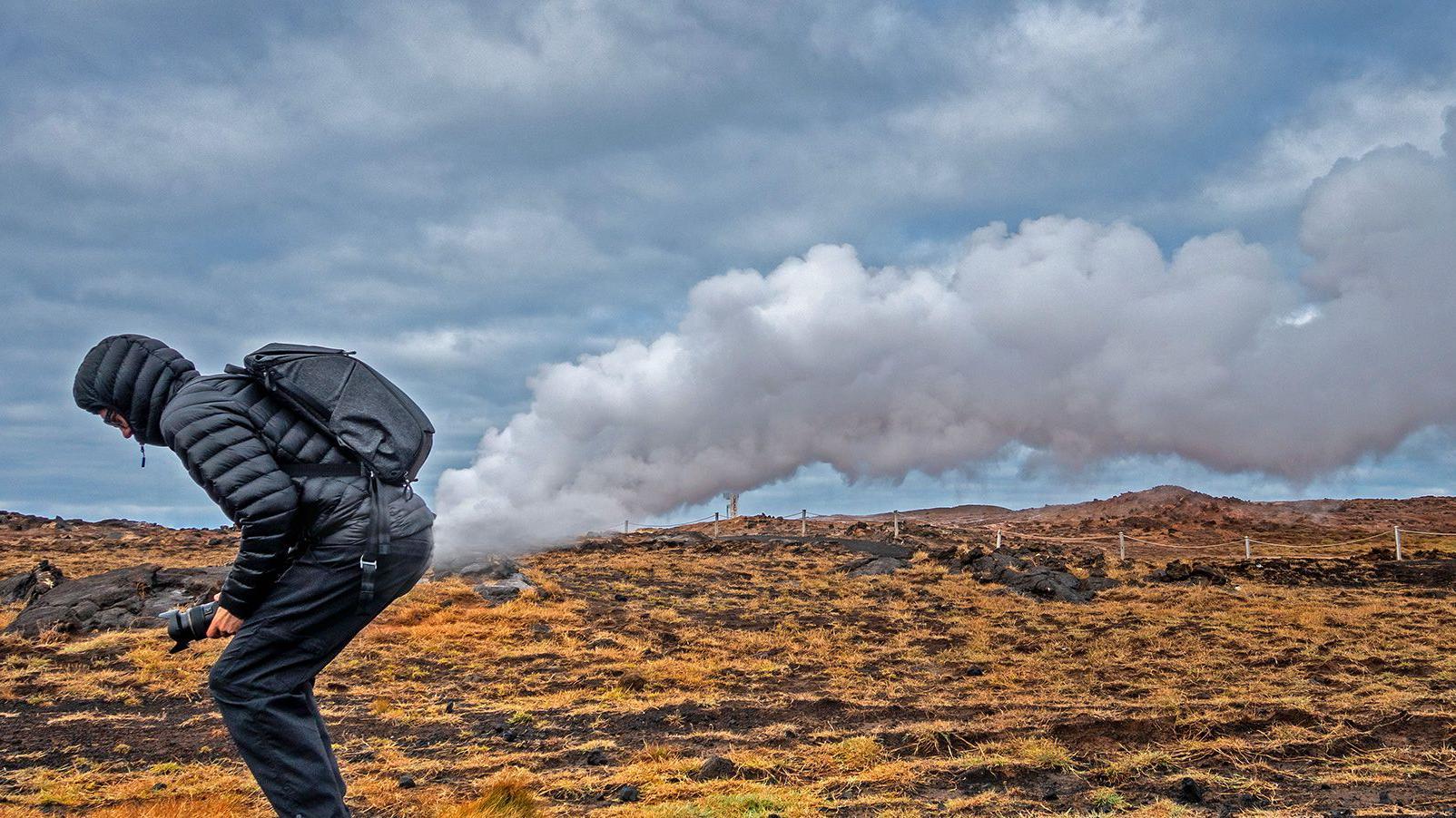 Stock image showing a man in a short puffer coat with a hood up, wearing walking trousers and shoes. He is squatting forward and has positioned himself so a burst of cloud looks like it is coming from his bottom. He is on a road next to a grassy landscape.