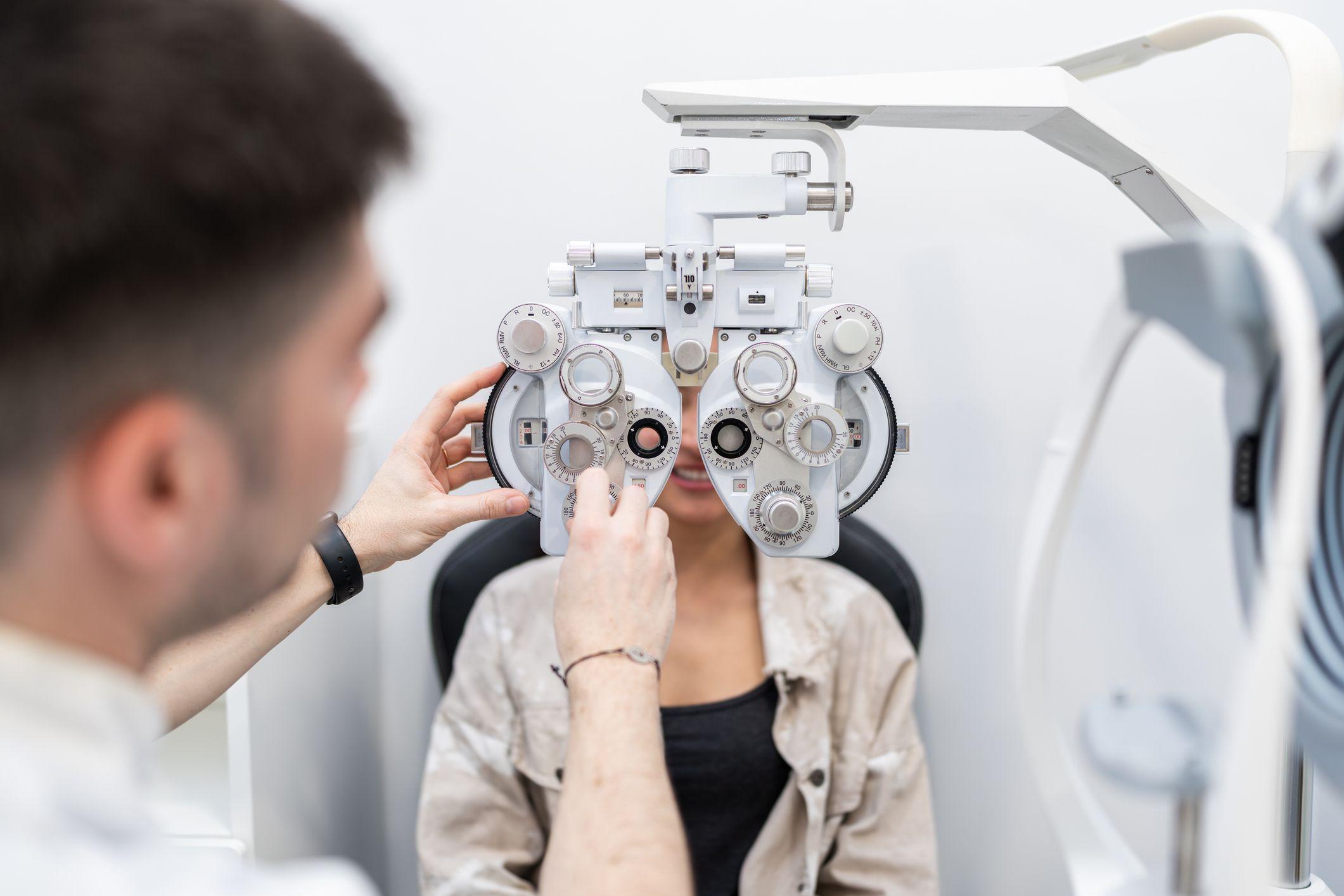 Una mujer está sentada en la silla del oftalmólogo y él le tiene el aparato para probar lentes en frente.