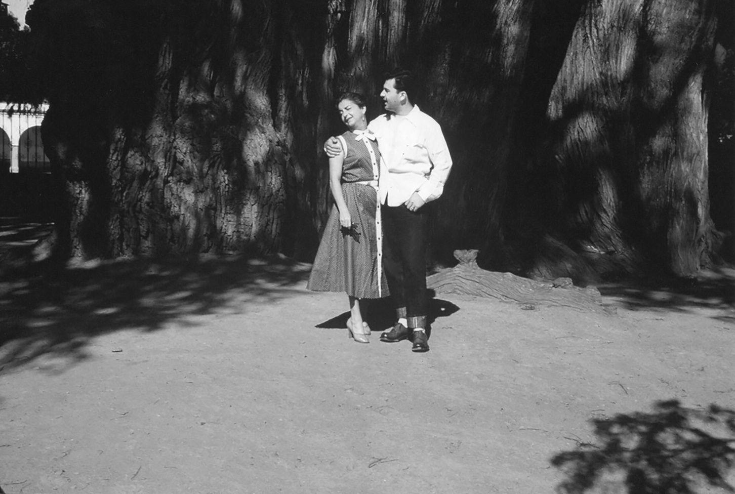Cristina y Antonio están parados abrazándose. Ella lleva un vestido largo sin mangas y tacones. Su hijo viste una camisa blanca y un pantalón oscuro. Están en lo que parece el patio de una casa en un día soleado.

