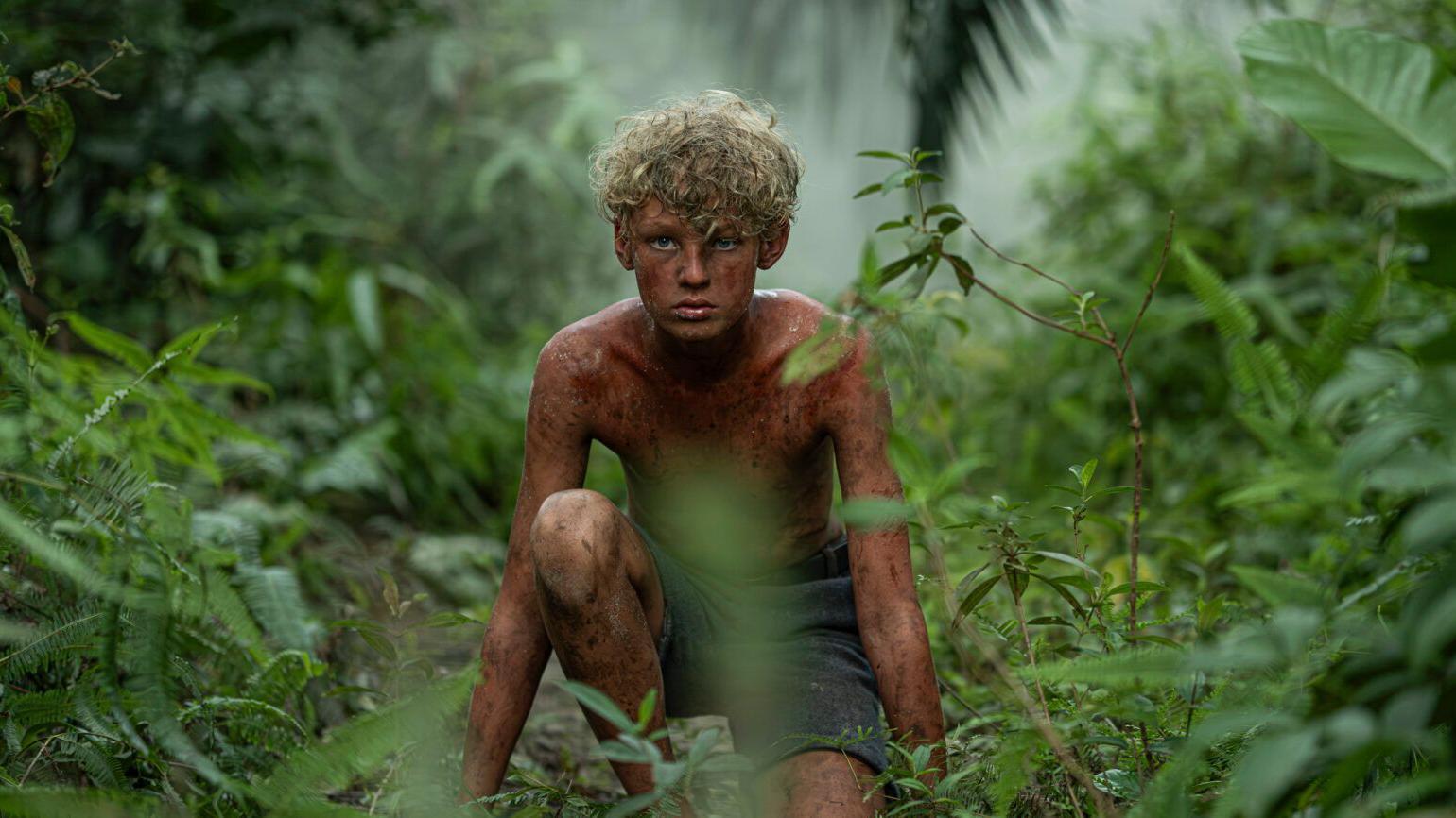 Un niño rubio en pantaloneta y sin camiseta en la selva. Está acurrucado, como si estuviera cazando. 