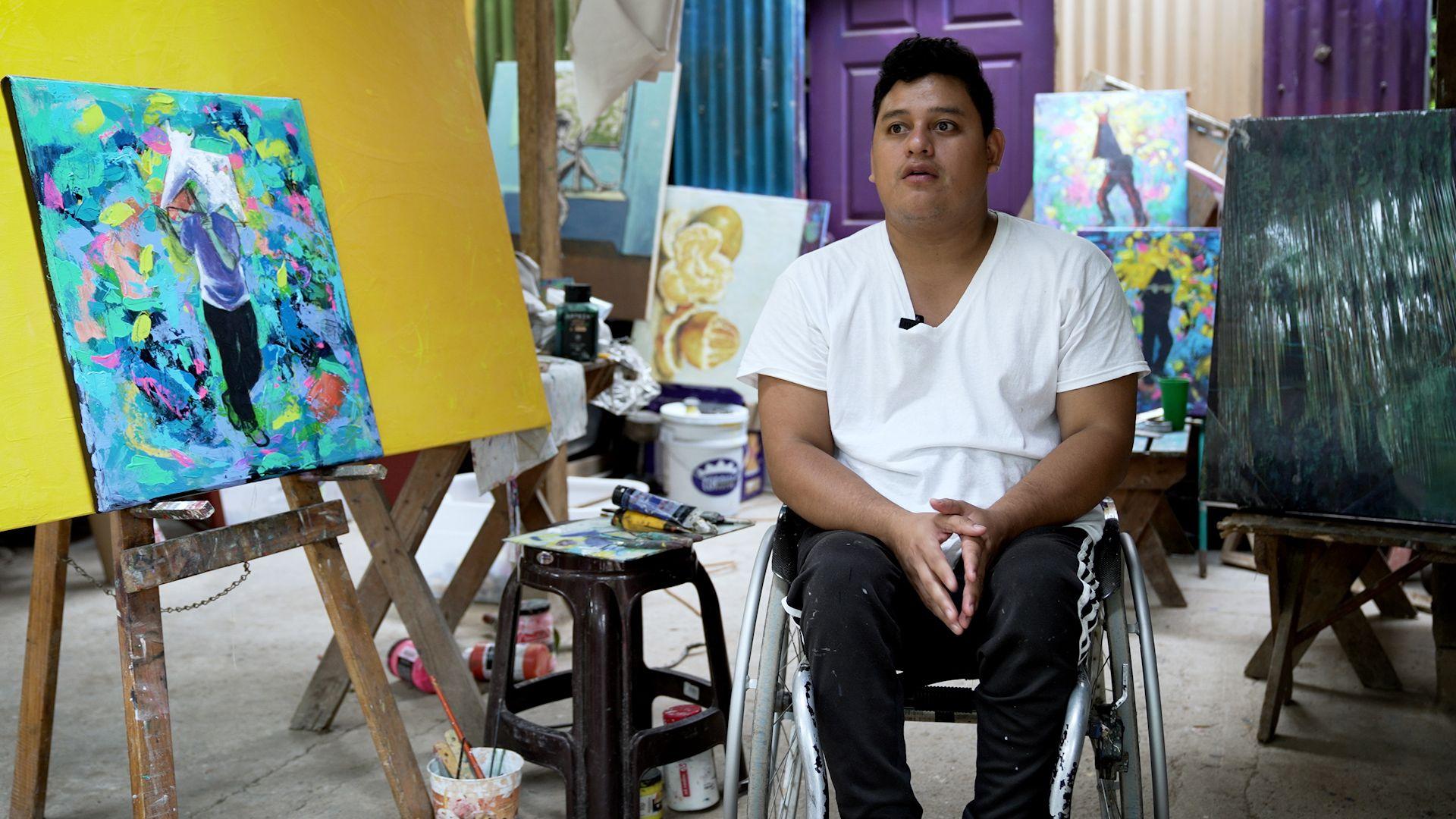 Homem em cadeira de rodas; ao lado dele, h&aacute; v&aacute;rias pinturas em cores vivas.