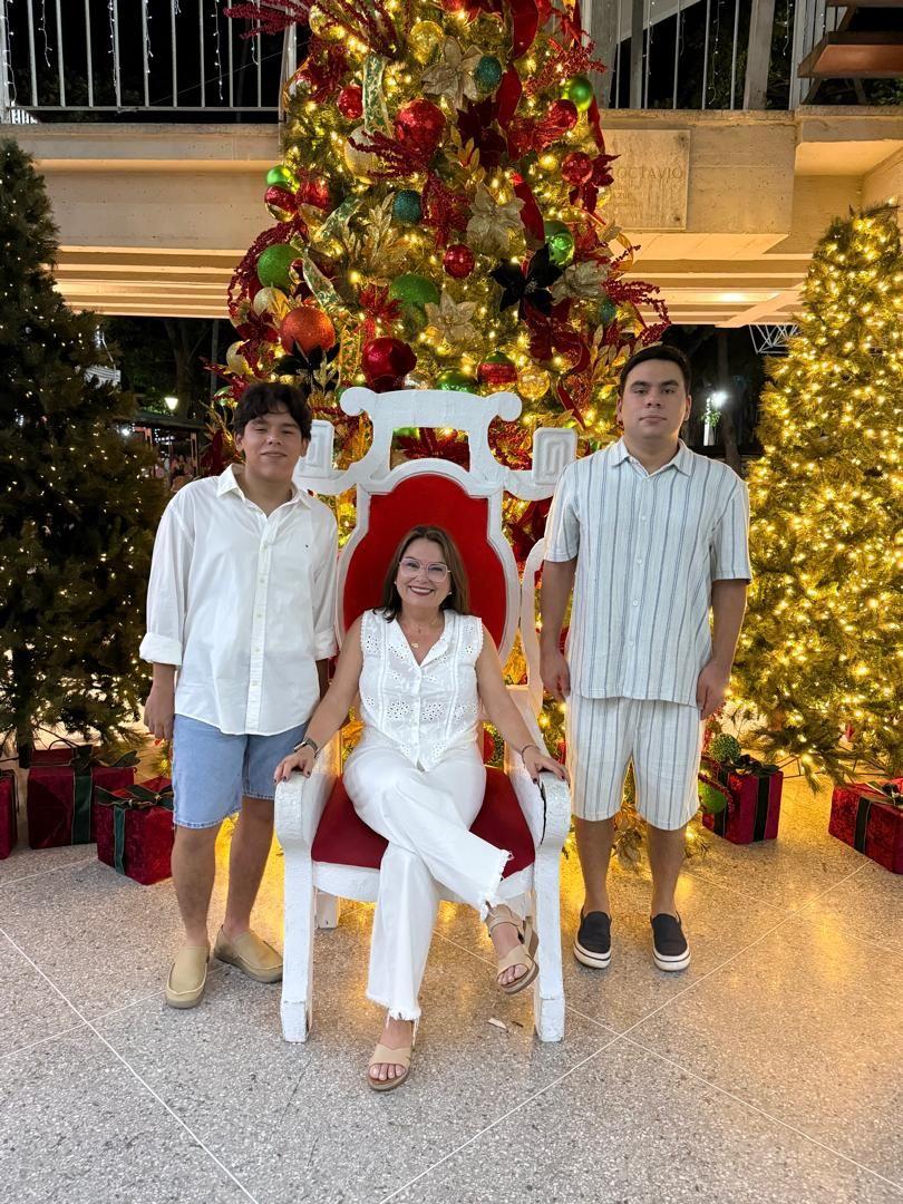 María Lourdes, vestida de blanco y con lentes, está sentada en una silla roja y blanca. A su lado derecho, está parado su hijo Gonzalo, que lleva una camisa blanca y un pantalón corto azul. A su lado izquierdo, está Santiago, que lleva un conjunto blanco con rayas oscuras. Al fondo tienen tres arboles de navidad decorados e iluminados.