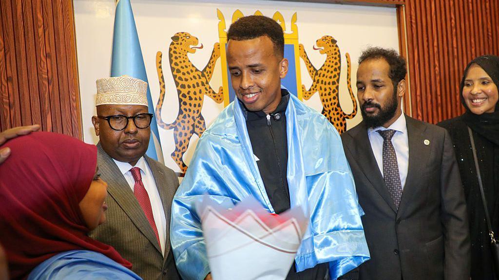 El árbitro somalí Omar Abdulkadir Artan, envuelto en la bandera somalí, recibe flores durante una ceremonia de bienvenida tras su llegada a Mogadiscio, Somalia, 9 de junio 2026