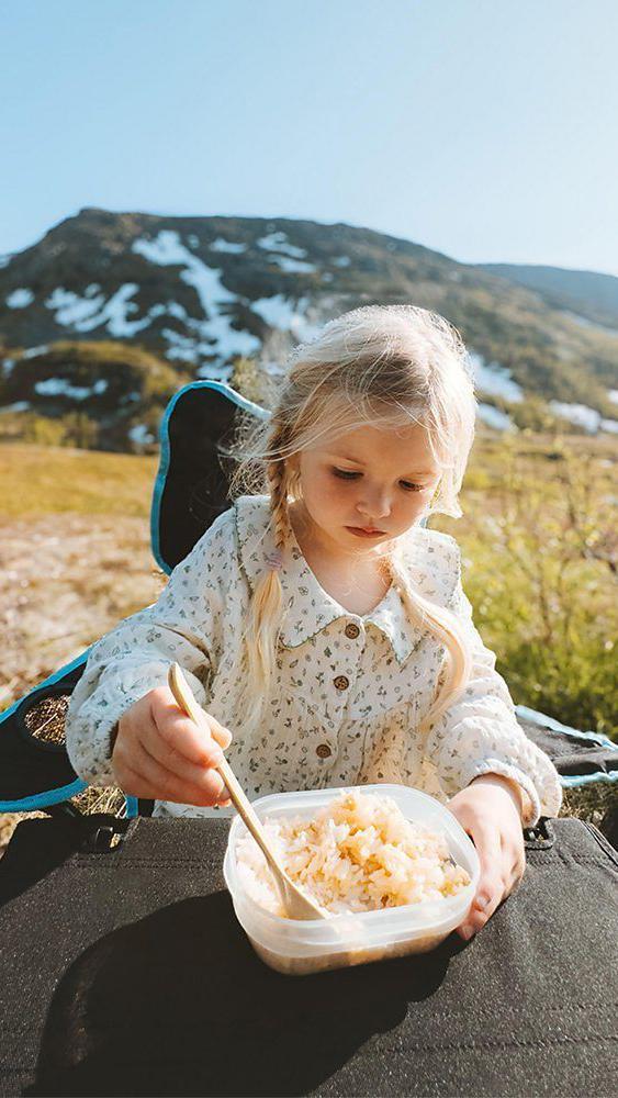 Menina come arroz em uma tigela plástica ao sol, em um campo aberto, com montanhas ao fundo
