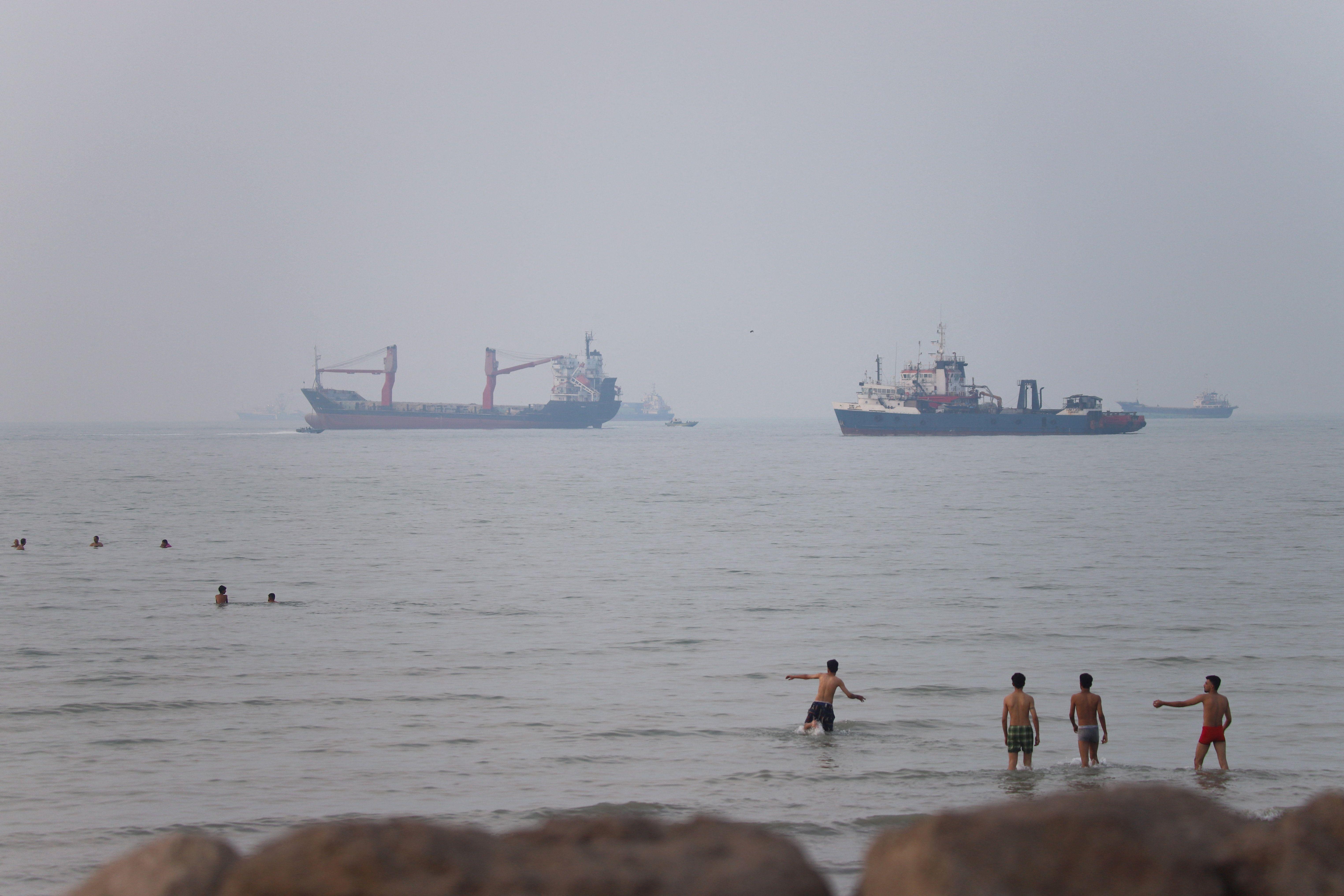 Buques de carga y petroleros permanecen en primer plano frente a la costa, mientras que personas nadan y se refrescan en las aguas costeras en un día brumoso.