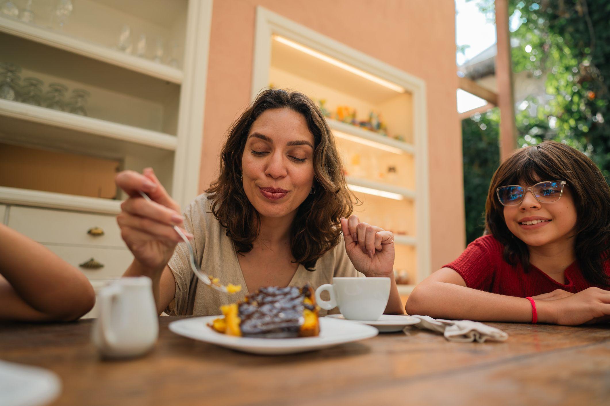 Mulher comendo um peda&ccedil;o de bolo de cenoura com calda de chocolate ao lado de uma menina de &oacute;culos