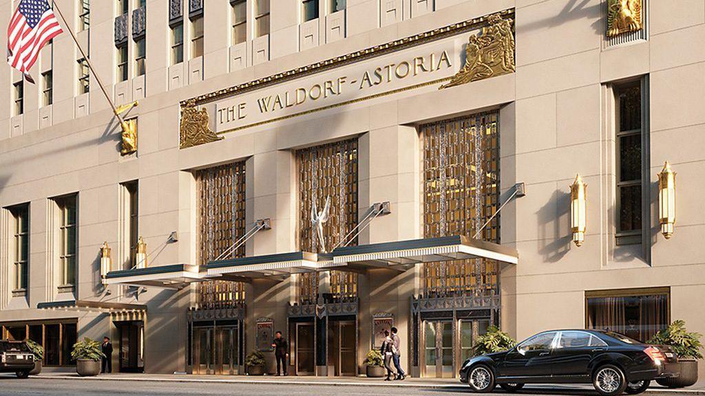 Fachada del Waldorf Astoria en Nueva York, de color arena con elementos dorados, y la bandera de Estados Unidos colgada de un mástil al costado del letrero donde se lee el nombre del hotel, y del otro lado una bandera negra con el logo del establecimiento en blanco.