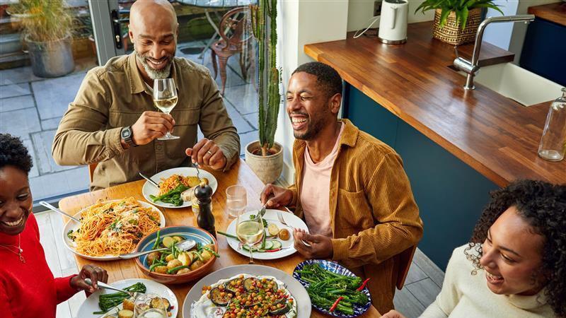Família rindo sentada em volta de uma mesa de madeira, comendo uma refeição. São servidos brócolis, batatas, azeitonas, peixe e berinjela.