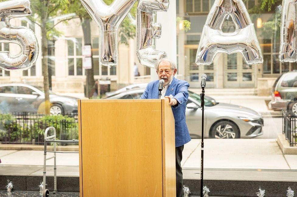 Marsel Mesulam en un atril hablando, detrás de él se pueden ver globos plateados que dicen 25 years (25 años)