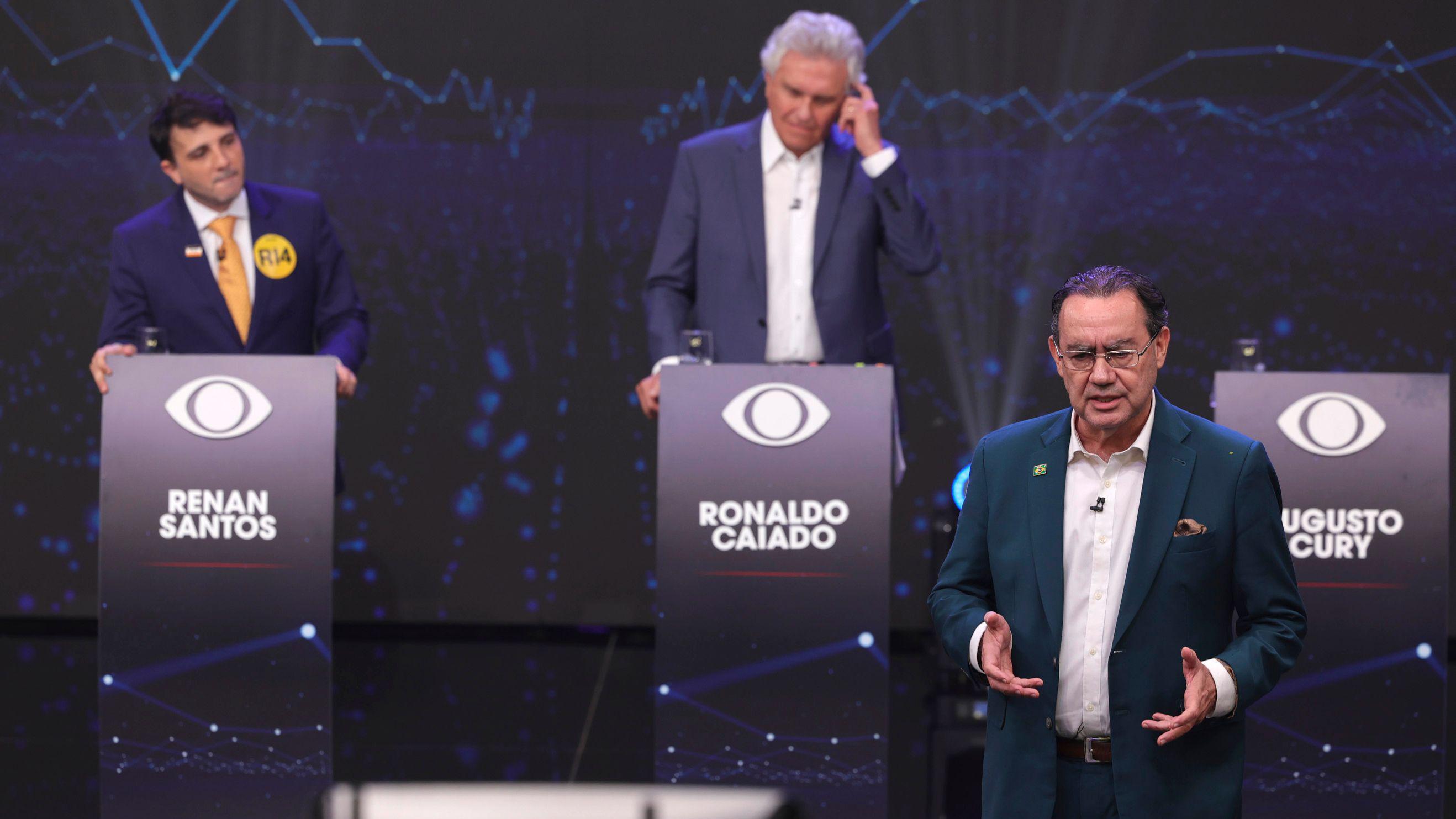Em um palco de debate, tr&ecirc;s candidatos aparecem diante de p&uacute;lpitos identificados com seus nomes. &Agrave; esquerda, Renan Santos observa a cena; ao centro, Ronaldo Caiado leva a m&atilde;o ao ouvido; e, em primeiro plano, Augusto Cury fala gesticulando. O fundo exibe gr&aacute;ficos em tons azulados e elementos visuais do evento.