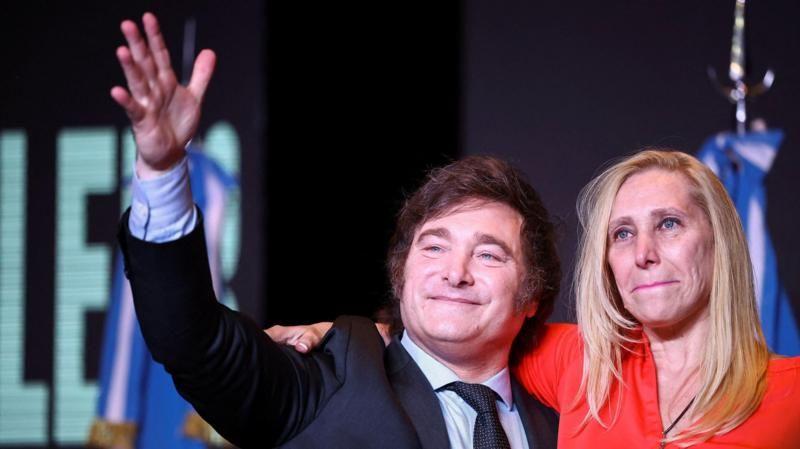 Javier e Karina Milei abraçados em evento; ele está acenando e sorrindo, ela mostra sorriso contido