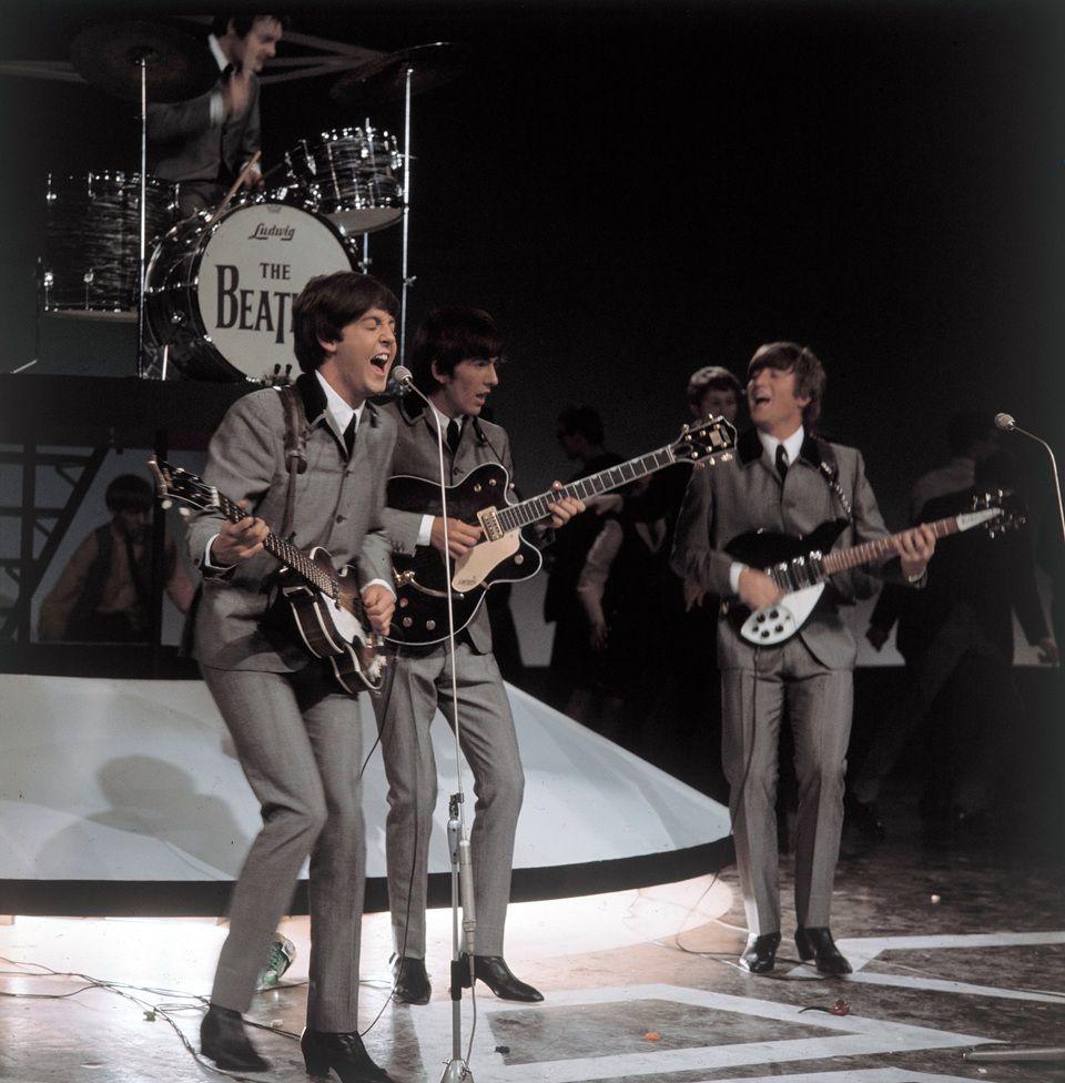 Paul McCartney toca la guitarra y canta frente a un micrófono. A su lado está George Harrison, también con su guitarra. A la izquierda está John Lennon. Atrás está la batería que dice The Beatles. Los cuatro miembros están vestidos igual con camisa blanca, corbata negra y chaqueta y pantalón grises. Están en una presentación

