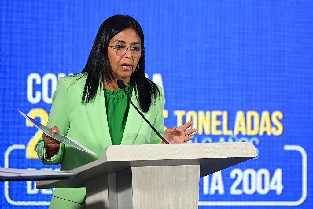 Delcy Rodr&iacute;guez, vestida com um traje de jaqueta verde, fala ao microfone em um p&uacute;lpito, com alguns pap&eacute;is na m&atilde;o.  