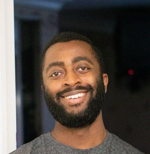 Abidemi Otaiku sorri para a c&acirc;mera. Ele &eacute; um homem negro, tem cabelo e barba preto, e est&aacute; em um ambiente interno, vestindo uma blusa cinza.