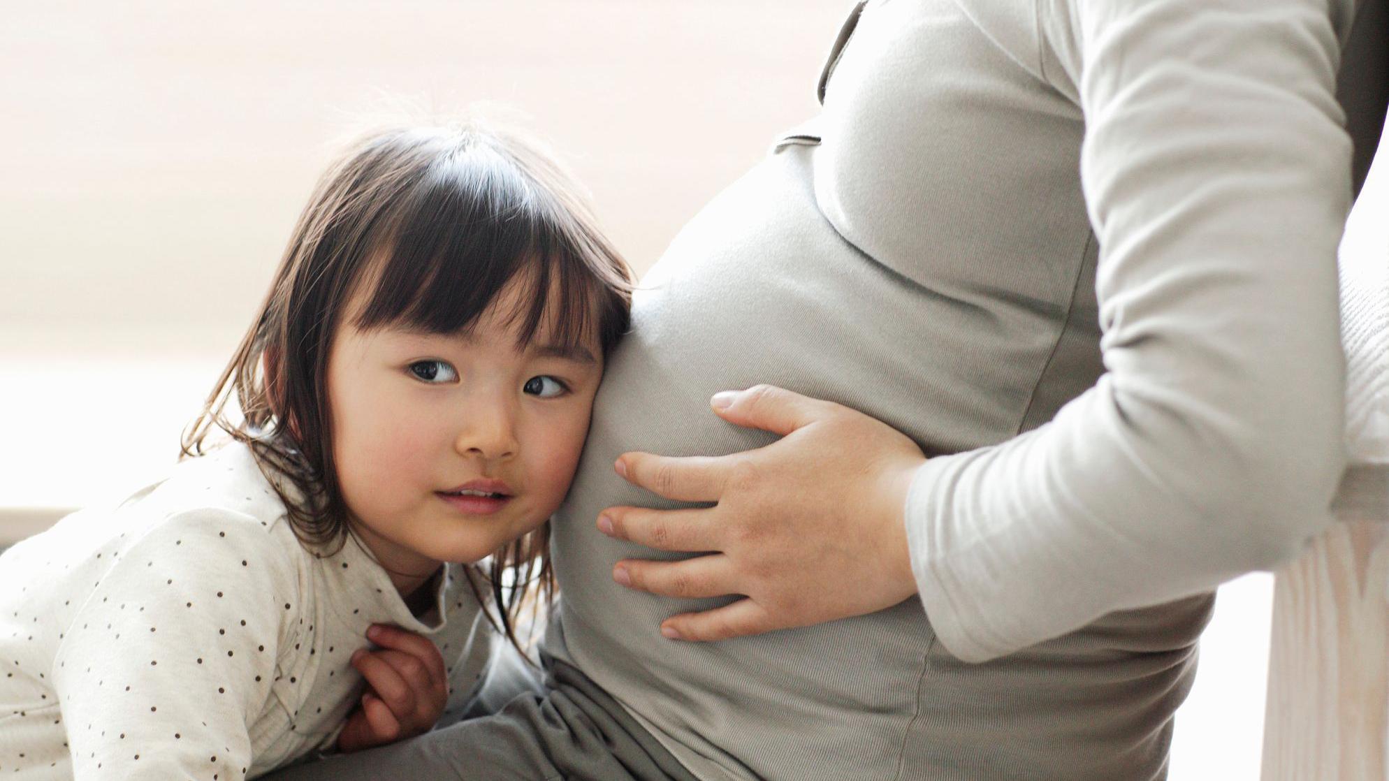 Una niña pequeña yace en el regazo de su madre, quien está en avanzado estado de gestación y se sujeta el vientre. La niña tiene la oreja pegada al vientre, escuchando los latidos del corazón.