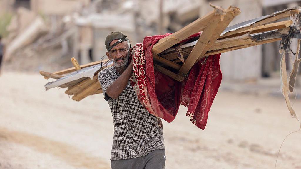 "أسوأ من البدء من الصفر": ما حجم مهمة إعادة إعمار غزة؟ 1 رجل يحمل حطباً بين الأنقاض في خان يونس، جنوب غزة، في 12 أكتوبر/تشرين الأول 2025، بينما تعود العائلات لاستعادة ممتلكاتها والتعامل مع الدمار الهائل الذي أعقب الانسحاب الإسرائيلي التدريجي.