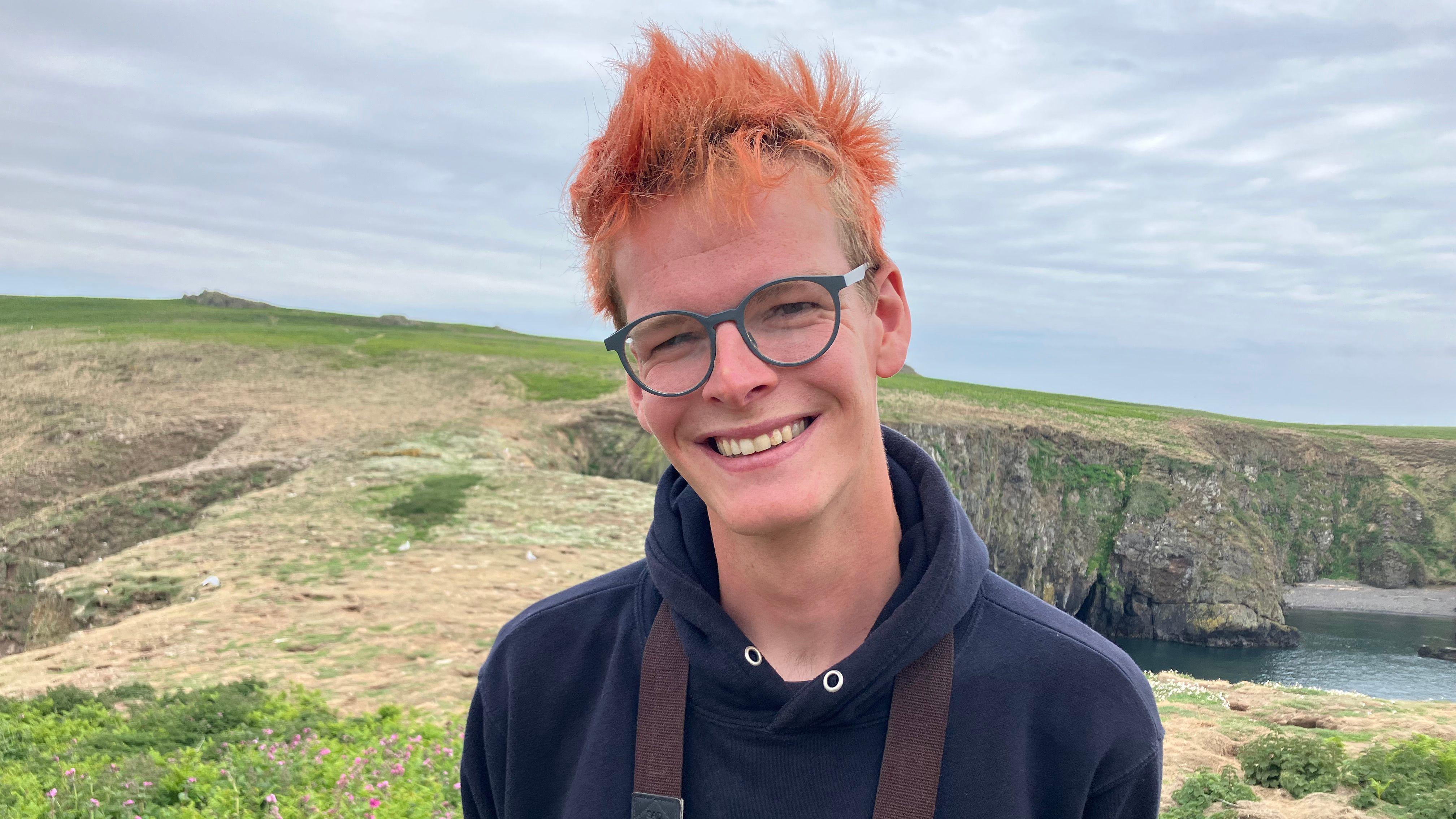 Un hombre mira a la cámara y sonríe. Está de pie en la isla Skomer con el mar a sus espaldas. Lleva una sudadera azul y gafas. Sonríe a la cámara y tiene el pelo pelirrojo.