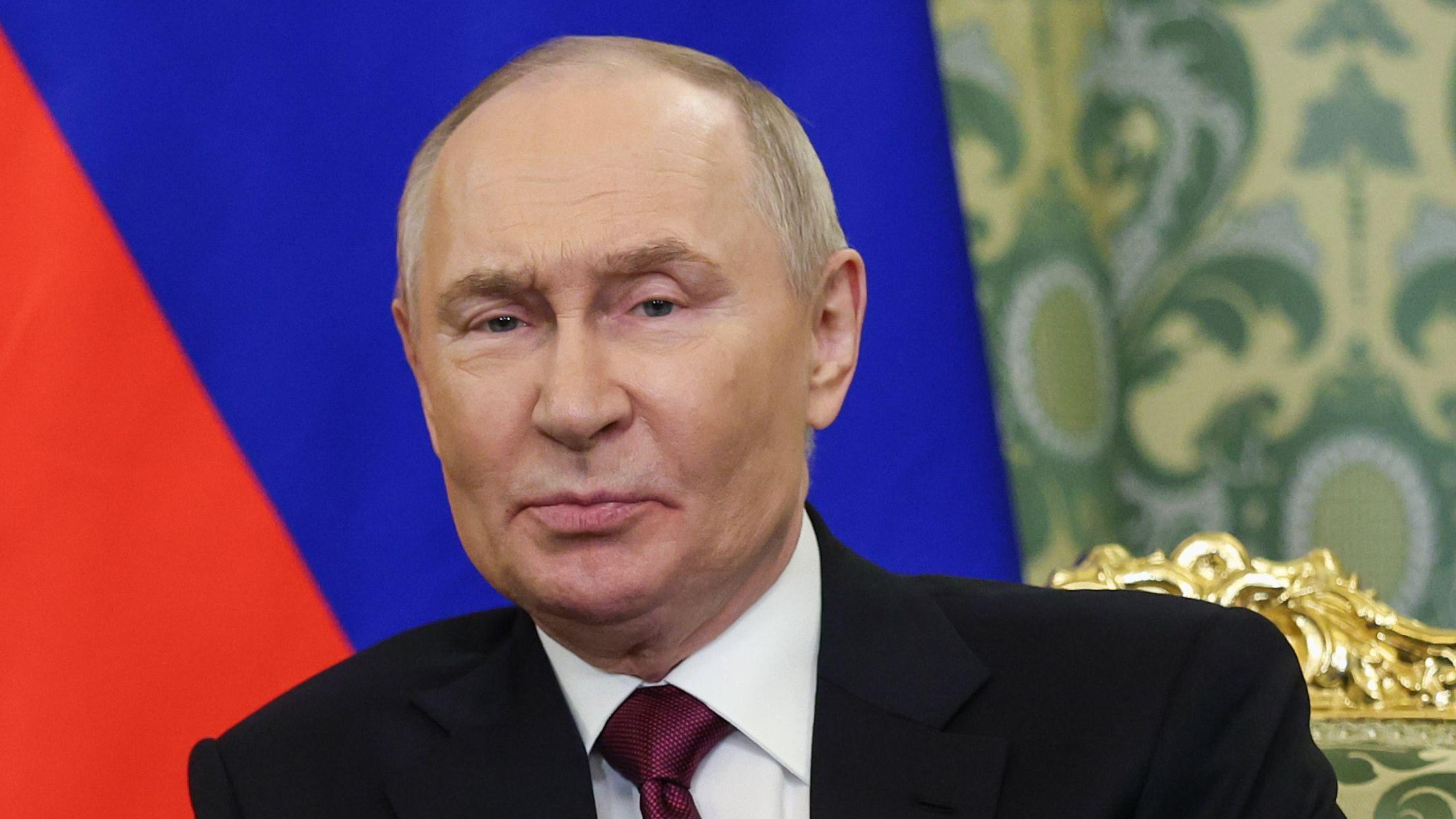 El presidente ruso Vladimir Putin está sentado en una silla frente a un telón de fondo con la bandera rusa.